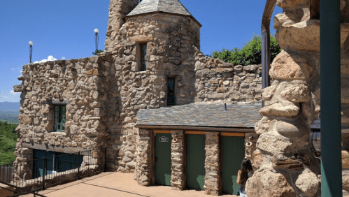 A stone building with a tower and green doors, surrounded by greenery and a clear blue sky.