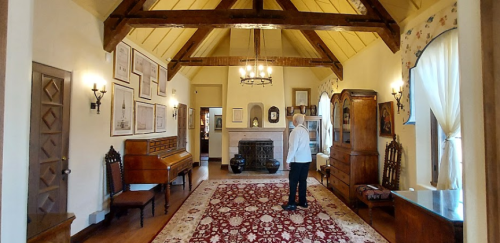 A spacious room with wooden beams, a large rug, antique furniture, and a person standing near a fireplace.