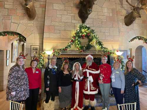 A group of nine people, including Santa and a woman in a red dress, pose by a festive fireplace decorated for Christmas.