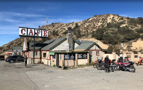 A rustic café building with a sign, surrounded by motorcycles and hills in the background.