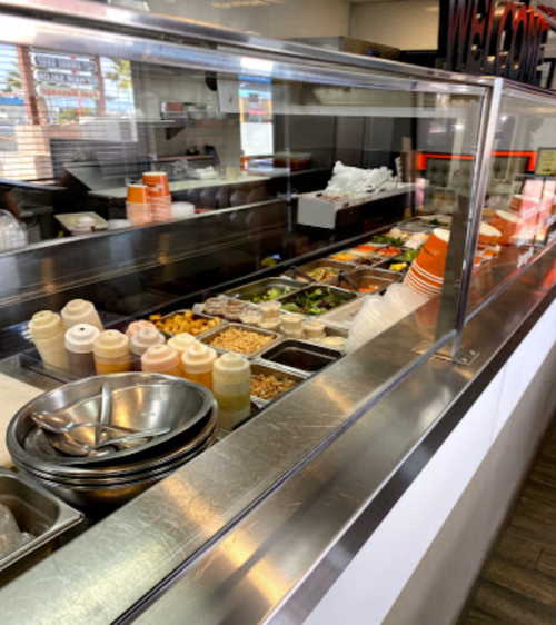 A food counter with various toppings, sauces, and fresh ingredients displayed behind a glass partition.