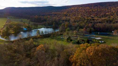 Aerial view of a serene landscape with a pond, trees in autumn colors, and rolling hills in the background.