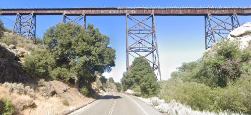A tall, rusted railway trestle bridge spans over a road surrounded by trees and rocky terrain under a clear blue sky.