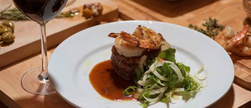 A gourmet dish featuring a steak topped with shrimp, served with greens and a glass of red wine in the background.