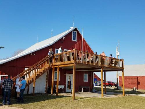 A red barn with a wooden deck, people gathered on the deck and around the entrance, under a clear blue sky.
