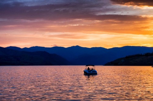 A boat floats on a calm lake at sunset, with mountains silhouetted against a colorful sky.
