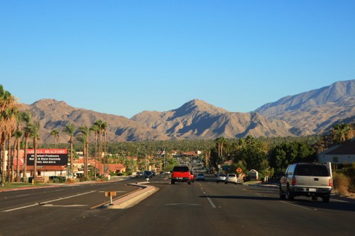 A scenic road view with mountains in the background, lined with palm trees and vehicles on the street.