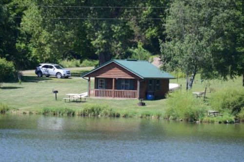 A small wooden cabin with a green roof by a pond, surrounded by trees and picnic tables, with a car parked nearby.