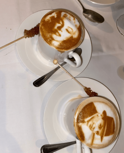Two cups of coffee with intricate foam art, featuring portraits, served on white plates with spoons and skewers.