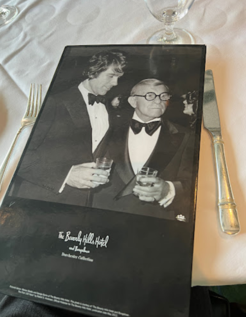 Black and white photo of two men in tuxedos holding drinks, with a menu from the Beverly Hills Hotel on the table.