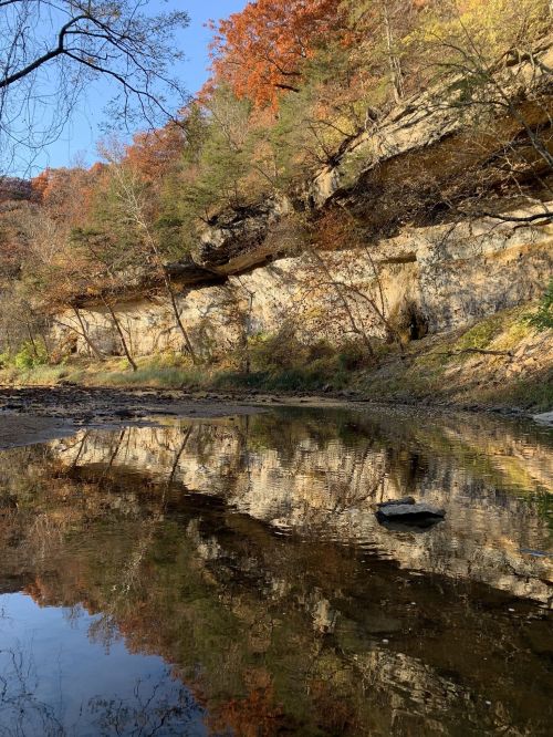 A serene riverbank scene with autumn foliage reflecting on calm water and rocky cliffs in the background.