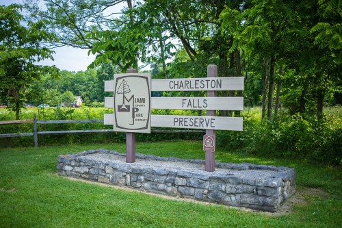 Sign for Charleston Falls Preserve, featuring a logo and wooden posts, surrounded by greenery and a stone base.