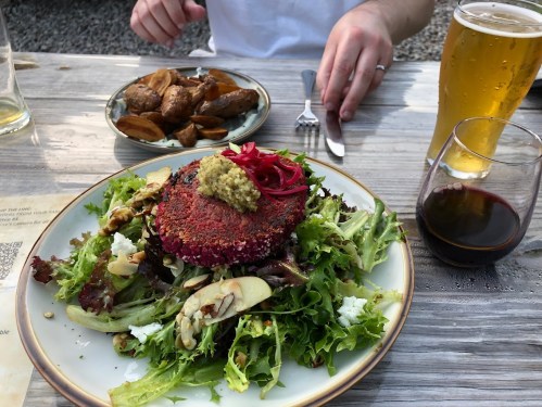 A colorful salad with a beet patty, topped with mustard and pickled onions, alongside roasted potatoes and drinks.