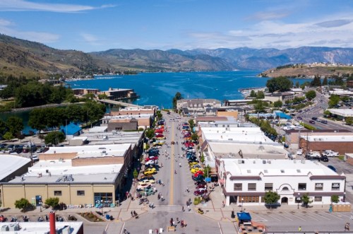 Aerial view of a vibrant lakeside town with shops, cars, and mountains in the background under a clear blue sky.