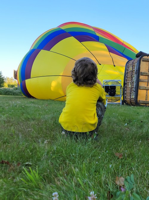 A child in a yellow shirt sits on the grass, watching a colorful hot air balloon being inflated.