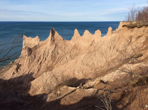 A rugged sandy cliff with sharp peaks overlooks a calm blue lake under a clear sky.