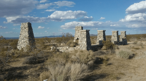 Ruins of stone structures in a desert landscape, surrounded by sparse vegetation and under a blue sky with clouds.