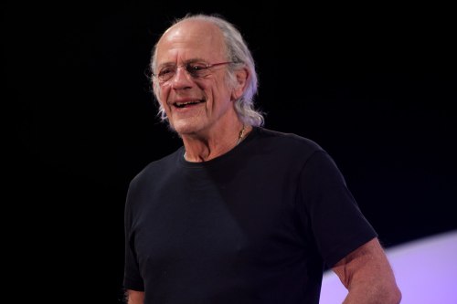 A smiling older man with long hair and glasses, wearing a black shirt, stands on stage against a dark background.