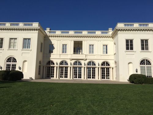 A large white mansion with arched windows and a balcony, set against a clear blue sky and green lawn.