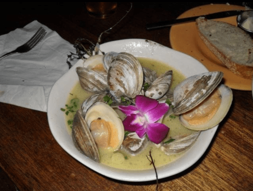 A bowl of clams in broth, garnished with an orchid, served with a slice of bread on the side.