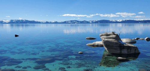 A serene lake scene with clear blue water, rocky formations, and distant snow-capped mountains under a bright sky.