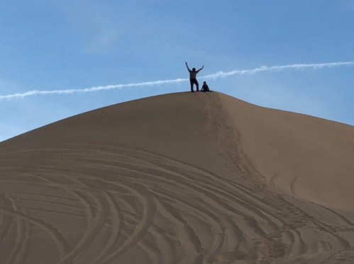 Two people stand on top of a sand dune, one with arms raised, against a blue sky with a trail of clouds.