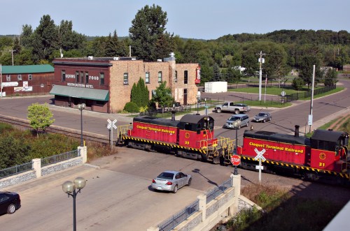 Two red locomotives cross a street near a brick building and parked cars, with trees in the background.