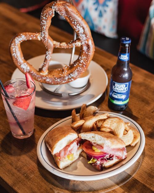 A large pretzel on a stand, a sandwich with toppings, fries, and a Bud Light beer on a wooden table.