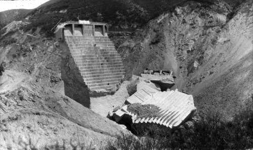 A partially collapsed concrete structure on a hillside, surrounded by rocky terrain and vegetation.