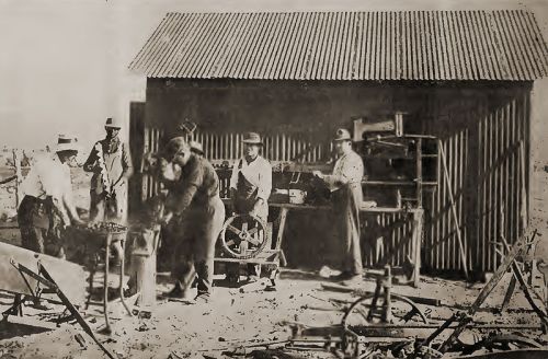 Black and white photo of workers in a workshop, using tools and machinery outside a shed.