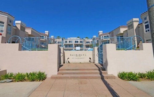 Entrance to a residential complex named "San Miguel," featuring blue railings and landscaped greenery under a clear blue sky.