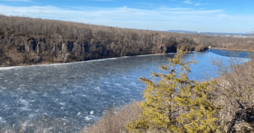 A scenic view of a frozen river surrounded by bare trees and rocky cliffs under a clear blue sky.
