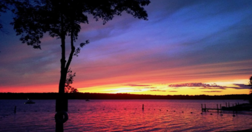 A serene lake at sunset, with vibrant orange and purple skies, silhouetted trees, and calm water reflecting the colors.