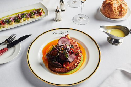 A gourmet dish featuring octopus, garnished with vegetables, served at a fine dining table with wine and bread.