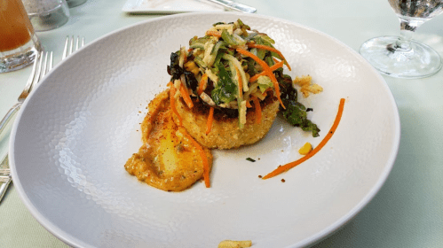A plated dish featuring a crispy round patty topped with shredded vegetables and a drizzle of sauce on a white plate.