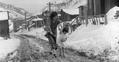 A woman carries a child and walks with a girl on a snowy, muddy road lined with small buildings.