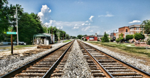 A view of railway tracks stretching into the distance, flanked by greenery and buildings under a blue sky.