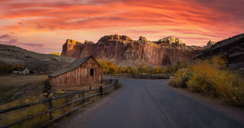 A rustic barn beside a winding road, with vibrant sunset colors illuminating rocky cliffs in the background.
