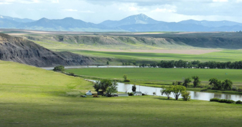 A scenic landscape featuring rolling green hills, a winding river, and distant mountains under a partly cloudy sky.