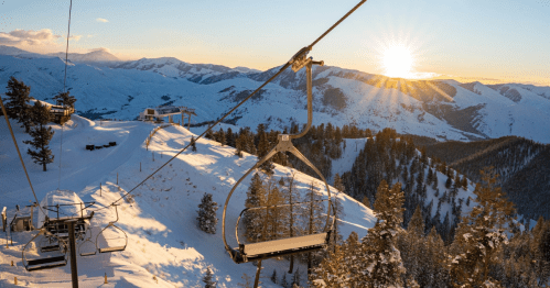 A ski lift overlooking snow-covered mountains at sunset, with trees in the foreground and a warm glow in the sky.