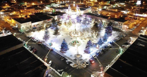 Aerial view of a festive town square illuminated with lights, decorated trees, and a carousel during the holiday season.