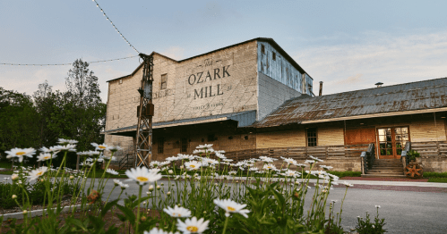 Historic Ozark Mill building with a rustic facade, surrounded by blooming daisies and a serene evening sky.