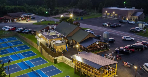 Aerial view of a sports complex with tennis courts, a restaurant, and parked cars in a rural area at dusk.