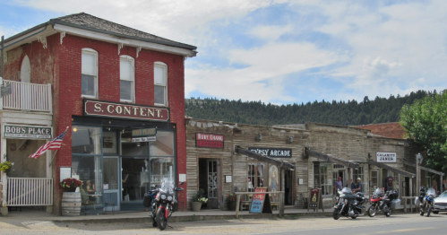 Historic buildings line a street in a small town, featuring shops and parked motorcycles under a cloudy sky.