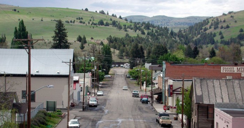 A quiet street lined with buildings and trees, set against rolling green hills under a cloudy sky.