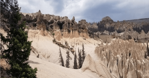 Rocky formations rise from sandy terrain under a cloudy sky, with sparse trees scattered throughout the landscape.