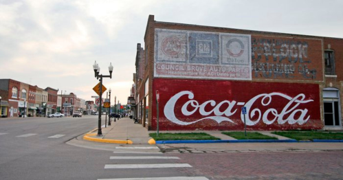 A street view featuring a Coca-Cola mural on a brick wall, with shops and a cloudy sky in the background.