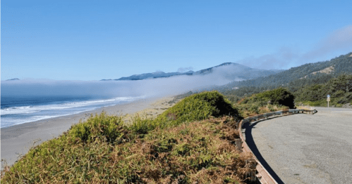 A scenic coastal view with fog rolling over the mountains and a sandy beach under a clear blue sky.