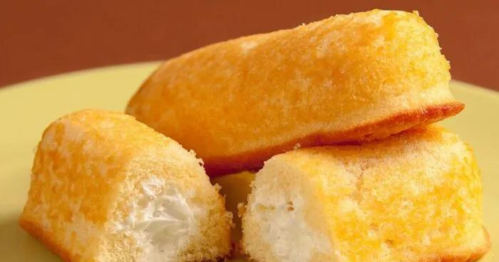 Three golden Twinkies, one cut in half to reveal creamy filling, on a yellow plate against a brown background.