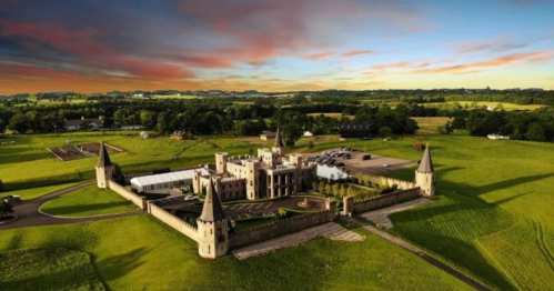 Aerial view of a castle surrounded by green fields and a colorful sunset sky.